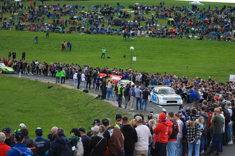 Zahlreiche Zuschauerinnen und Zuschauer säumen eine Straße bei einem Bergrennen. Entlang der Strecke stehen dicht gedrängt Menschen, während Rennfahrzeuge vorbeifahren, im Hintergrund sitzen weitere Besucher auf einer Wiese.