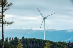 Eine Windkraftanlage auf einem bewaldeten Berg, im Hintergrund in höhergelegenen Almgelände mehr als zehn weitere Windräder