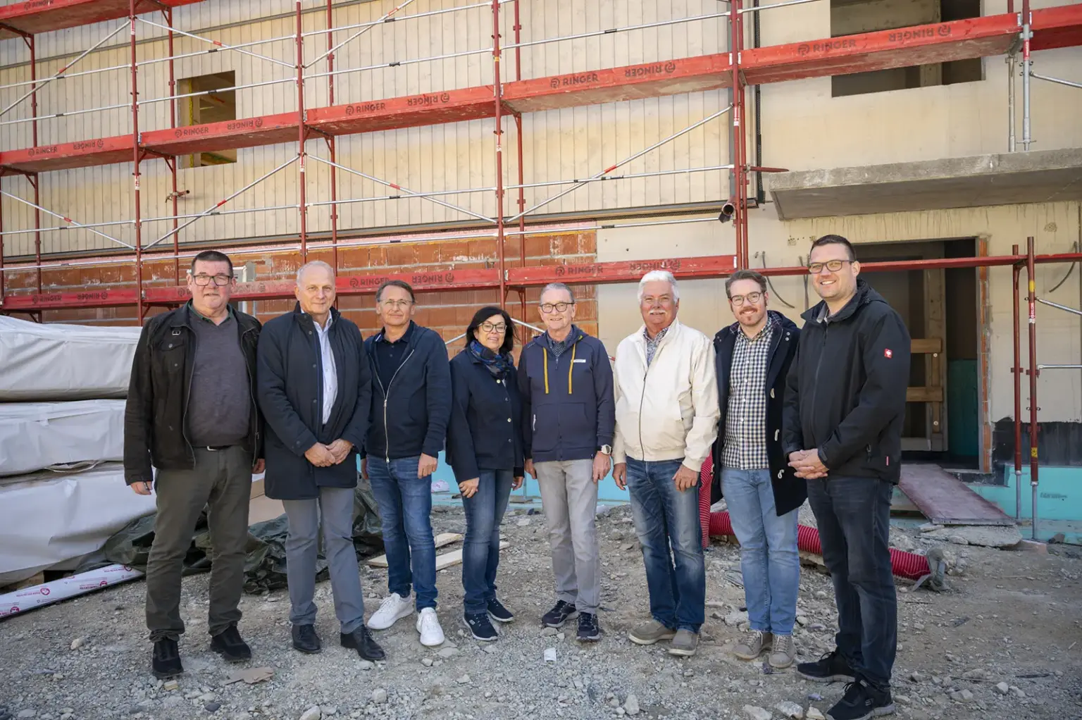 Eine Gruppe von acht Personen steht vor einem im Bau befindlichen Gebäude mit Gerüst. Die Beteiligten sind in Alltagskleidung gekleidet und posieren auf der Baustelle im Rahmen einer Gleichenfeier.