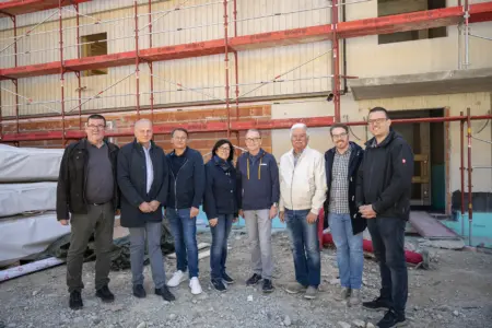 Eine Gruppe von acht Personen steht vor einem im Bau befindlichen Gebäude mit Gerüst. Die Beteiligten sind in Alltagskleidung gekleidet und posieren auf der Baustelle im Rahmen einer Gleichenfeier.