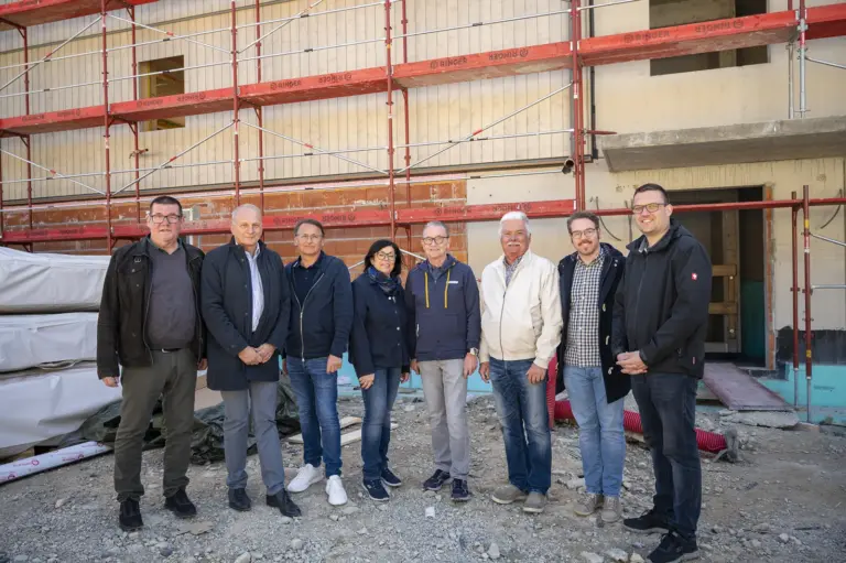 Eine Gruppe von acht Personen steht vor einem im Bau befindlichen Gebäude mit Gerüst. Die Beteiligten sind in Alltagskleidung gekleidet und posieren auf der Baustelle im Rahmen einer Gleichenfeier.