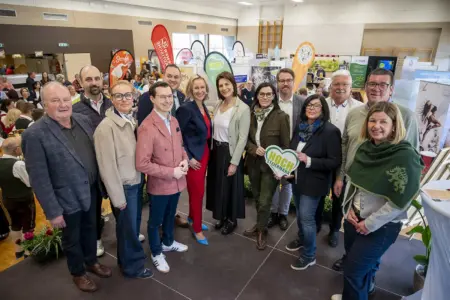 Eine Gruppe von rund einem Dutzend Erwachsenen steht in einer Messehalle vor Informationsständen und lächelt in die Kamera; im Hintergrund sind Besucher, Werbebanner und Ausstellerstände zu sehen.