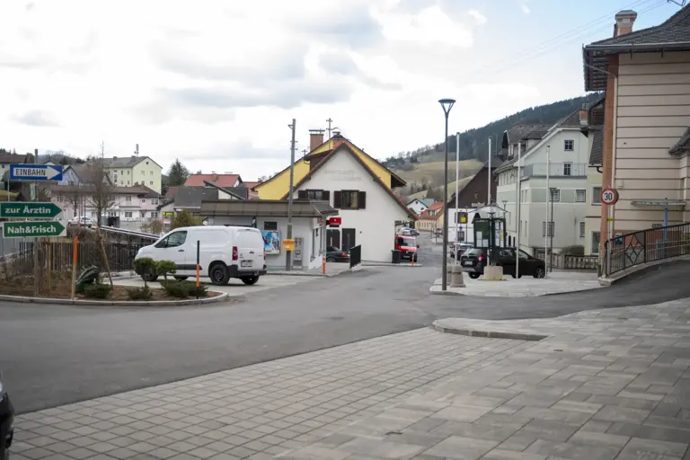 Ein neu gestalteter Ortsbereich mit Straßenkreuzung und Gehwegen im Ortszentrum. In der Mitte steht ein Gebäude mit Geschäft, daneben sind parkende Autos, eine Bushaltestelle und Wegweiser zu sehen, im Hintergrund Wohnhäuser und Hügel.