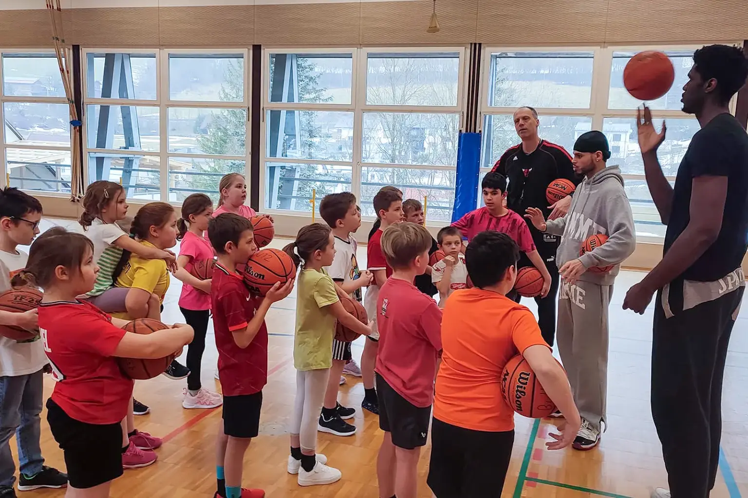Eine Gruppe von Kindern mit Basketbällen und drei große Männer stehen in einem Turnsaal.