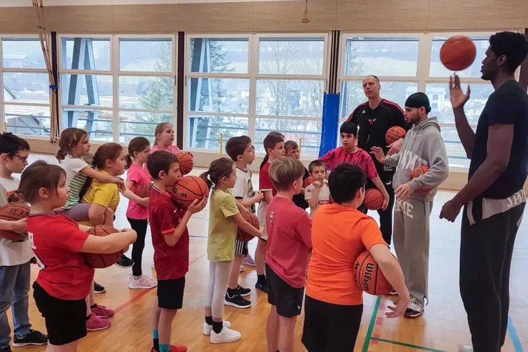 Eine Gruppe von Kindern mit Basketbällen und drei große Männer stehen in einem Turnsaal.