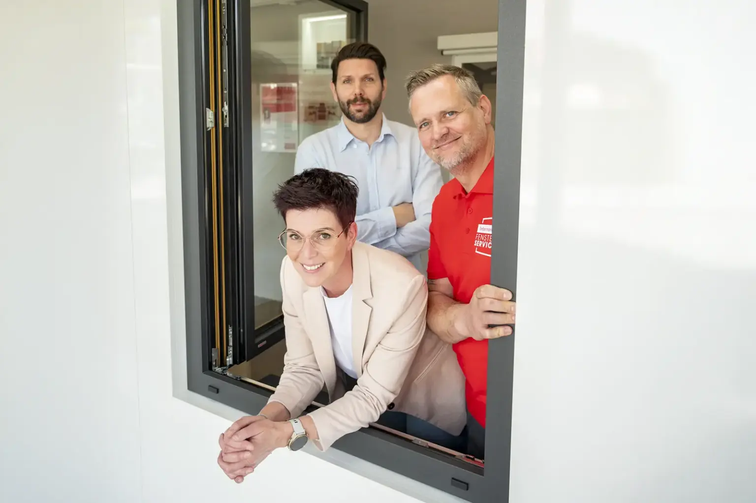 Drei Erwachsene schauen aus einem geöffneten Fenster eines modernen Gebäudes: vorne lehnt eine Frau im hellen Blazer am Rahmen, hinter ihr stehen zwei Männer, einer im Hemd, der andere in einem roten Shirt mit Firmenlogo.