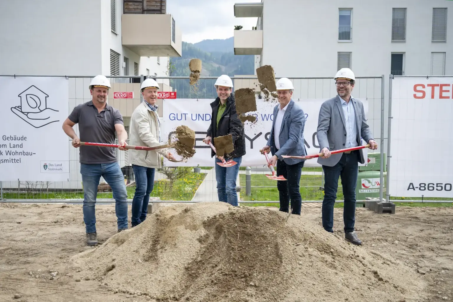 Spatenstich für neue Wohnungen in St. Marein Fünf Männer mit Bauhelmen führen auf einer Baustelle einen symbolischen Spatenstich durch. Sie werfen gleichzeitig Erde in die Luft, im Hintergrund sind Wohngebäude und Bautafeln zu sehen.