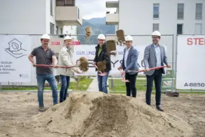 Fünf Männer mit Bauhelmen führen auf einer Baustelle einen symbolischen Spatenstich durch. Sie werfen gleichzeitig Erde in die Luft, im Hintergrund sind Wohngebäude und Bautafeln zu sehen.