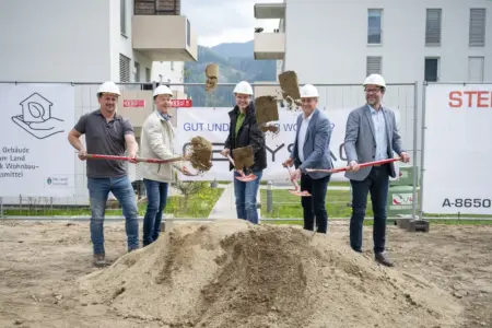 Fünf Männer mit Bauhelmen führen auf einer Baustelle einen symbolischen Spatenstich durch. Sie werfen gleichzeitig Erde in die Luft, im Hintergrund sind Wohngebäude und Bautafeln zu sehen.