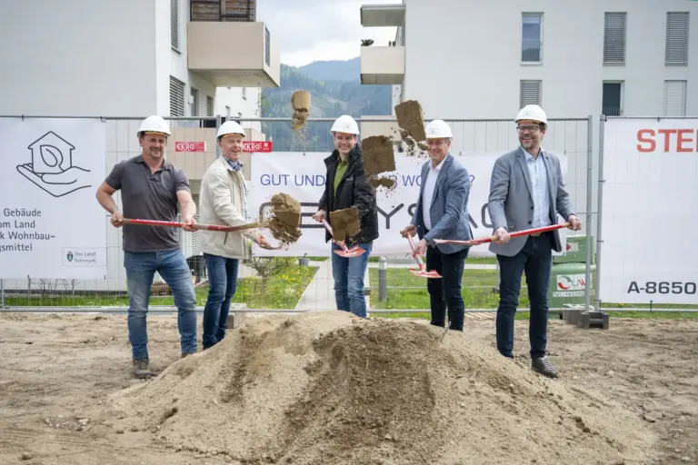 Fünf Männer mit Bauhelmen führen auf einer Baustelle einen symbolischen Spatenstich durch. Sie werfen gleichzeitig Erde in die Luft, im Hintergrund sind Wohngebäude und Bautafeln zu sehen.