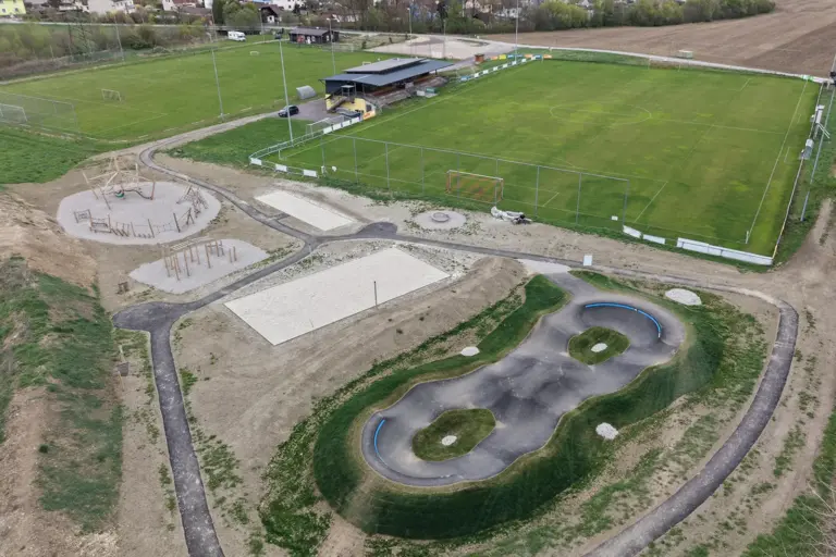 Luftaufnahme eines Freizeitgeländes mit asphaltierter Buckelbahn für Radfahrer, Beachvolleyball- und Bocciafeld sowie Klettergeräten, im Hintergrund zwei Fußballfelder und ein Kabinen- und Tribünengebäude