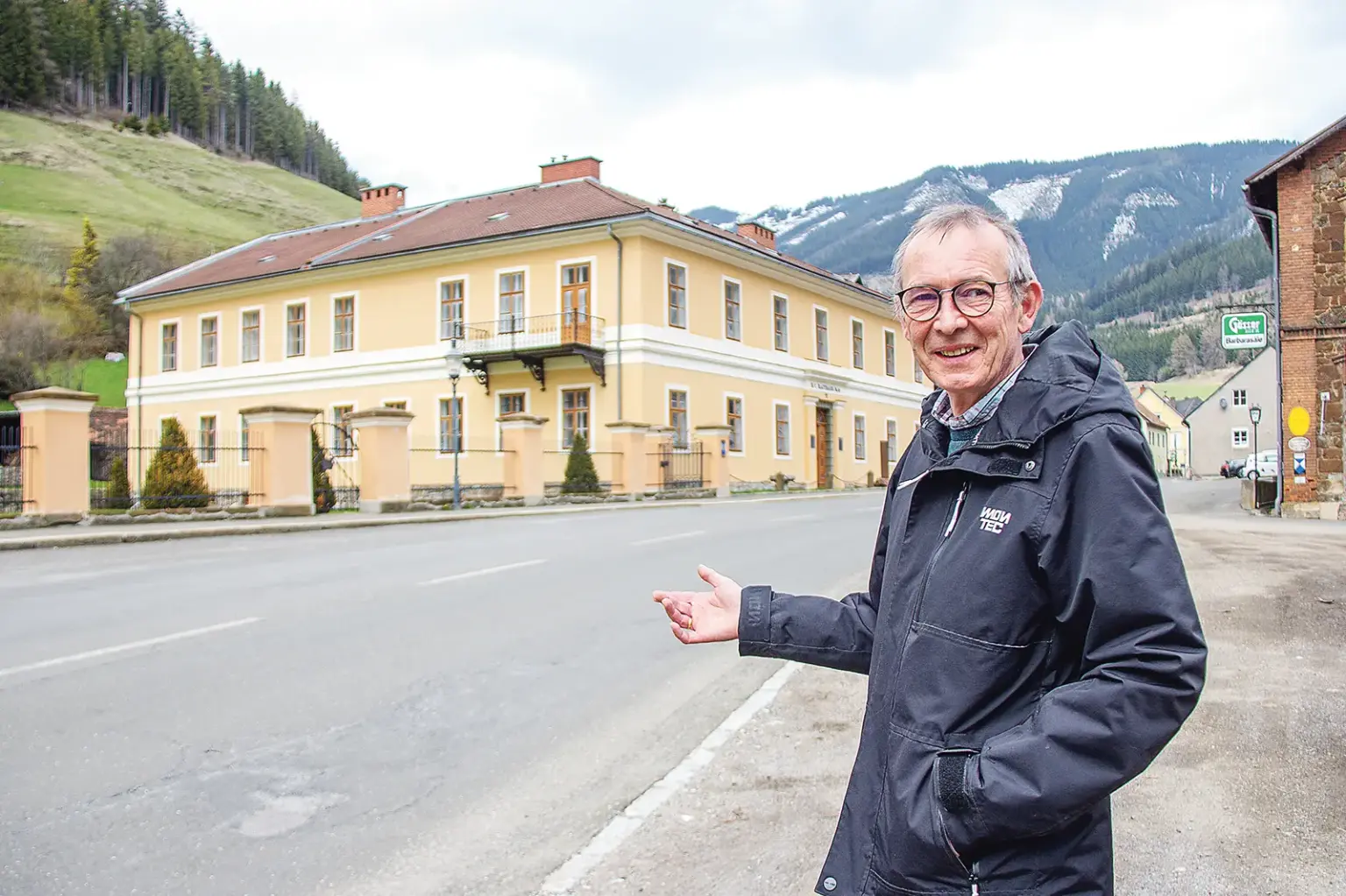 Ein Mann mit Brille steht neben einer Straße, im Hintergrund ein historisches Haus mit gelber Fassade