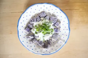 Afoch Guat: Frühlingskräuter auf dem Teller Von oben fotografierter Teller mit hausgemachten violetten Gnocchi, serviert mit cremiger Kräutersauce und frischen Frühlingszwiebeln. Das Gericht ist auf einem weiß-blau gepunkteten Teller angerichtet.