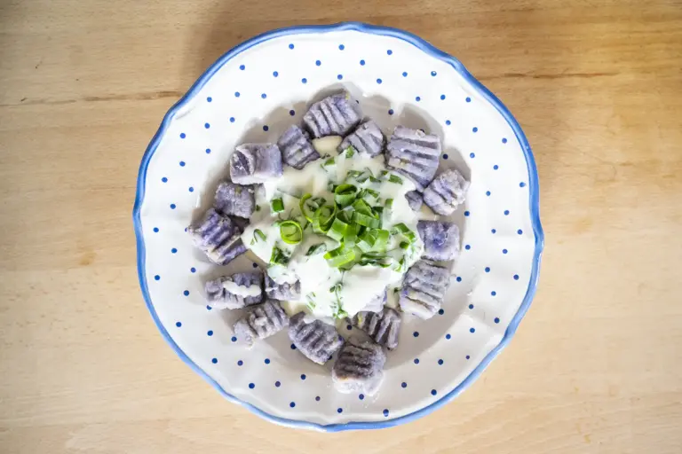 Von oben fotografierter Teller mit hausgemachten violetten Gnocchi, serviert mit cremiger Kräutersauce und frischen Frühlingszwiebeln. Das Gericht ist auf einem weiß-blau gepunkteten Teller angerichtet.