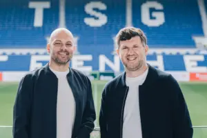 Zwei Männer stehen lächelnd im Stadion vor den blau-weißen Tribünen der TSG Hoffenheim. Das gemeinsame Porträt entstand am Spielfeldrand in sportlichem Umfeld.