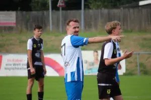 Ein Fußballspieler im blau-weißen Trikot zeigt während eines Spiels mit ausgestrecktem Arm über den Platz und gibt Anweisungen; um ihn herum stehen weitere Spieler auf dem Rasen und verfolgen die Spielsituation.