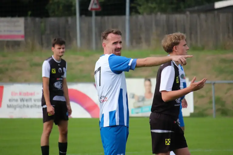 Ein Fußballspieler im blau-weißen Trikot zeigt während eines Spiels mit ausgestrecktem Arm über den Platz und gibt Anweisungen; um ihn herum stehen weitere Spieler auf dem Rasen und verfolgen die Spielsituation.