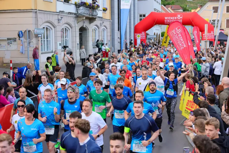 Sport und Party beim Brucker Businesslauf Menschen mit Startnummern laufen dicht gedrängt auf einer Straße, links und rechts Publikum