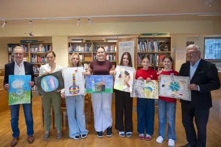 Acht Personen – darunter mehrere Schülerinnen – stehen in einer Schulbibliothek nebeneinander und präsentieren selbst gestaltete Bilder zum Thema Frieden. Links und rechts begleiten zwei Erwachsene die Gruppe, im Hintergrund sind Bücherregale und Lernbereiche zu sehen.