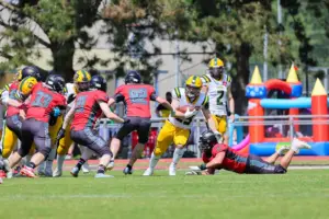 Spieler zweier American-Football-Teams kämpfen auf dem Spielfeld um Raumgewinn. Ein Ballträger in gelb-weißer Ausrüstung versucht durch die Verteidigung in roten Trikots zu brechen, während im Hintergrund eine Hüpfburg zu sehen ist.