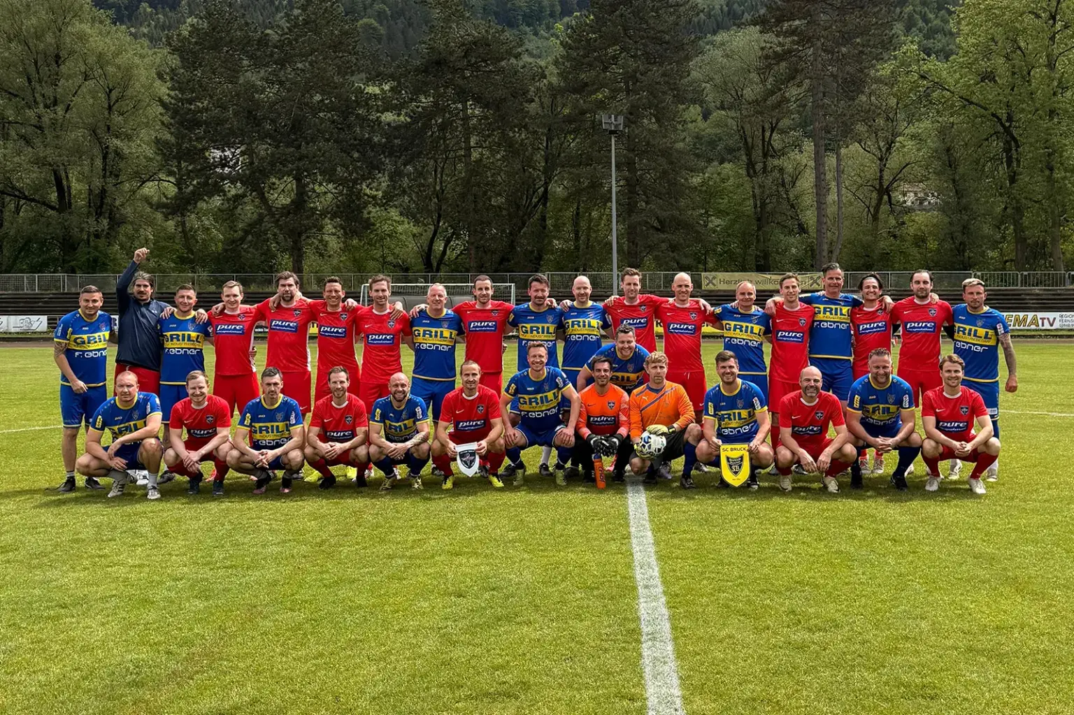 Benefizkick im Murinselstadion Gruppenfoto auf einem Fußballplatz: Spieler zweier Mannschaften in roten und blau-gelben Trikots posieren vor einem Benefizspiel gemeinsam auf dem Rasen vor bewaldeter Kulisse.