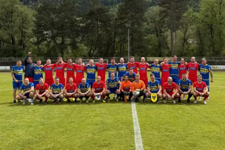 Gruppenfoto auf einem Fußballplatz: Spieler zweier Mannschaften in roten und blau-gelben Trikots posieren vor einem Benefizspiel gemeinsam auf dem Rasen vor bewaldeter Kulisse.