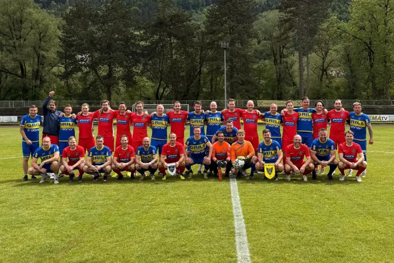Gruppenfoto auf einem Fußballplatz: Spieler zweier Mannschaften in roten und blau-gelben Trikots posieren vor einem Benefizspiel gemeinsam auf dem Rasen vor bewaldeter Kulisse.