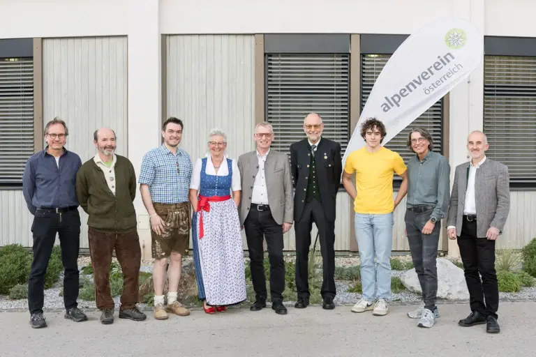 Eine Frau und acht Männer vor einem Gebäude und einer Fahne mit Logo des Alpenvereins.