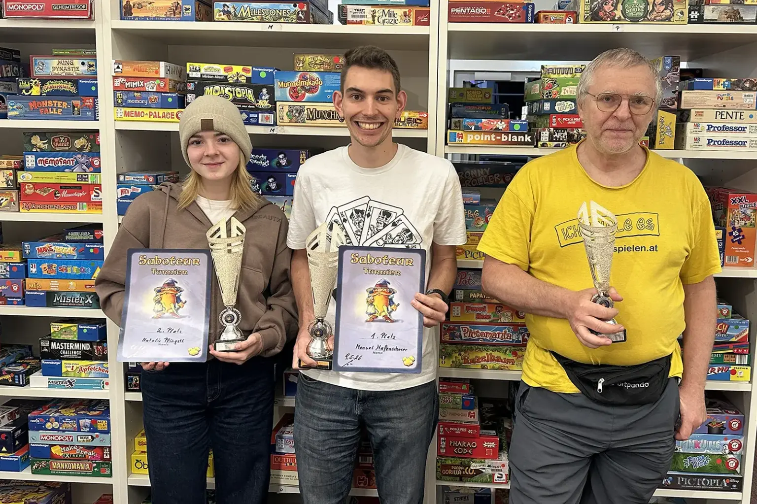 „Saboteur“- Meisterschaft mit Brucker Doppelsieg Eine Frau und zwei Männer mit Pokal und Urkunden. Im Hintergrund eine Regalwand mit Brettspielen.