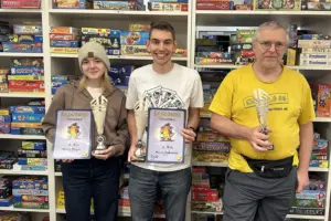 Eine Frau und zwei Männer mit Pokal und Urkunden. Im Hintergrund eine Regalwand mit Brettspielen.