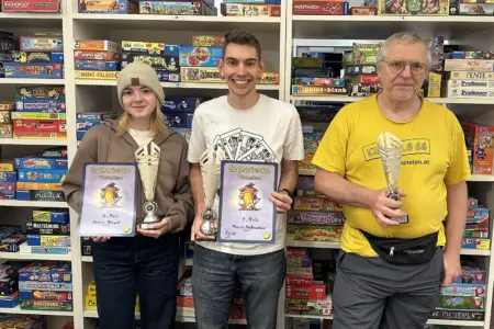 Eine Frau und zwei Männer mit Pokal und Urkunden. Im Hintergrund eine Regalwand mit Brettspielen.