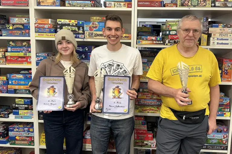 Eine Frau und zwei Männer mit Pokal und Urkunden. Im Hintergrund eine Regalwand mit Brettspielen.