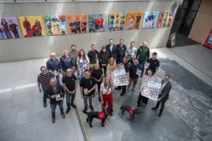 Die Stadt Bruck lädt zur ersten Vereinsmesse Rund 25 Vertreterinnen und Vertreter verschiedener Vereine präsentieren in einer Ausstellungshalle Flyer und Plakate der Vereinsmesse. Im Vordergrund stehen zwei Rettungshunde, im Hintergrund hängen farbenfrohe Porträts.