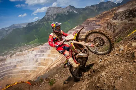 Ein Motorradfahrer fährt beim Erzbergrodeo mit seiner Enduro steil einen Schotterhang hinauf. Im Hintergrund sind der terrassenförmige Erzberg und die umliegende Berglandschaft unter blauem Himmel zu sehen.