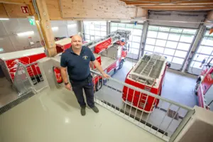 Ein Mann in Feuerwehrkleidung steht an einer Brüstung in einer Halle mit großen Ausfahrtstoren und Feuerwehrfahrzeugen