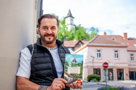 Ein Mann mit Buch in Händen steht auf einem innerstädtischen Platz, im Hintergrund ein Turm auf einer Anhöhe
