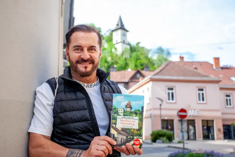 Verlosung: Fünf neue Preitler-Krimis zu gewinnen Ein Mann mit Buch in Händen steht auf einem innerstädtischen Platz, im Hintergrund ein Turm auf einer Anhöhe