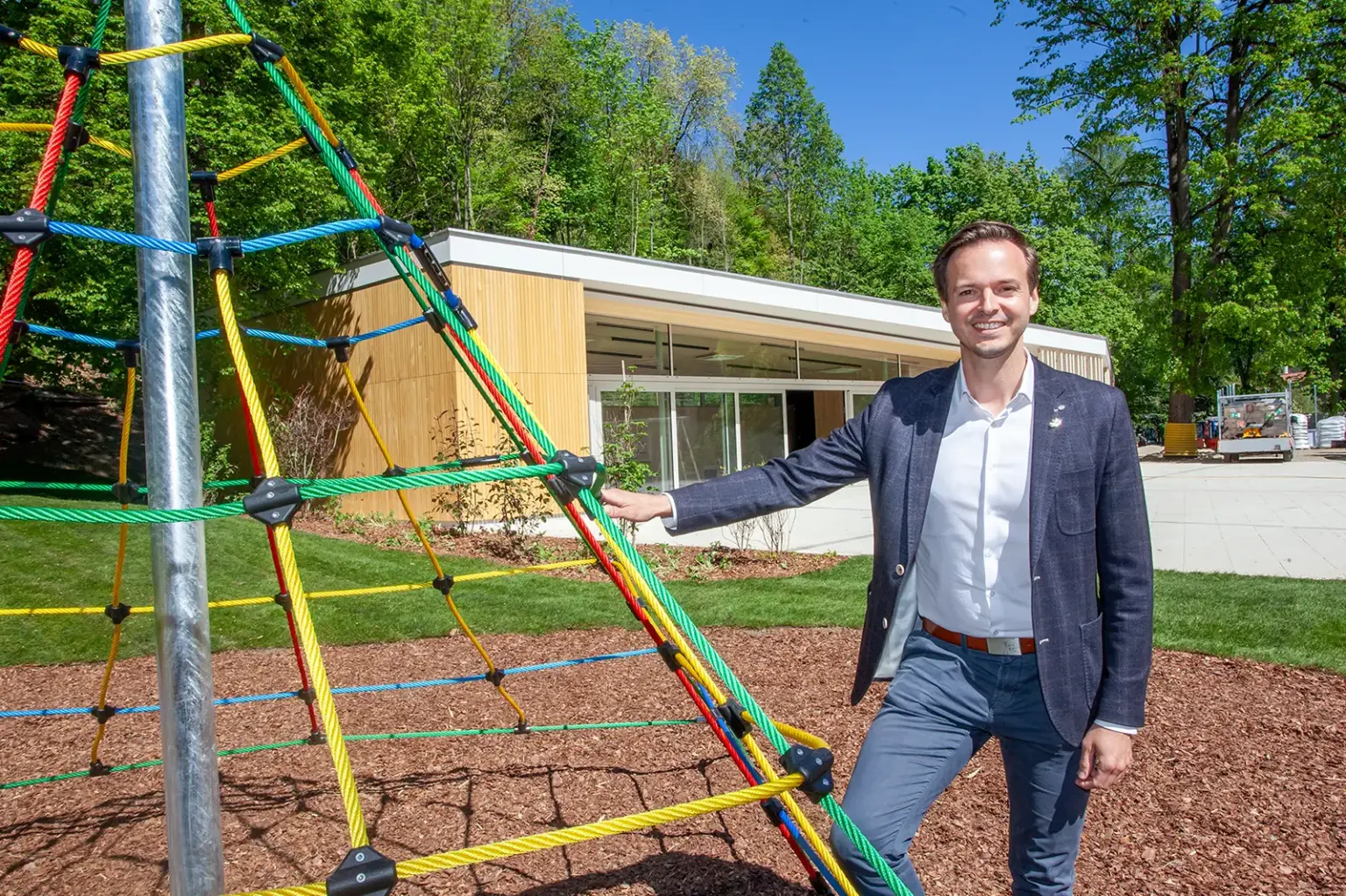 Ein Mann steht an einem Kletter-Spielgerät, im Hintergrund ein Holzbau und zahlreiche Bäume
