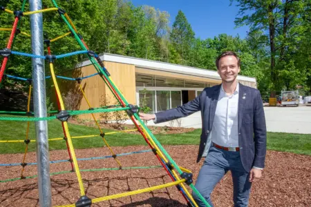 Ein Mann steht an einem Kletter-Spielgerät, im Hintergrund ein Holzbau und zahlreiche Bäume
