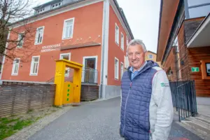 Ein Mann steht lächelnd auf der Straße, im Hintergrund links ein Haus mit der Aufschrift "Volksschule Kindergarten", rechts ein Haus mit Holzfassade