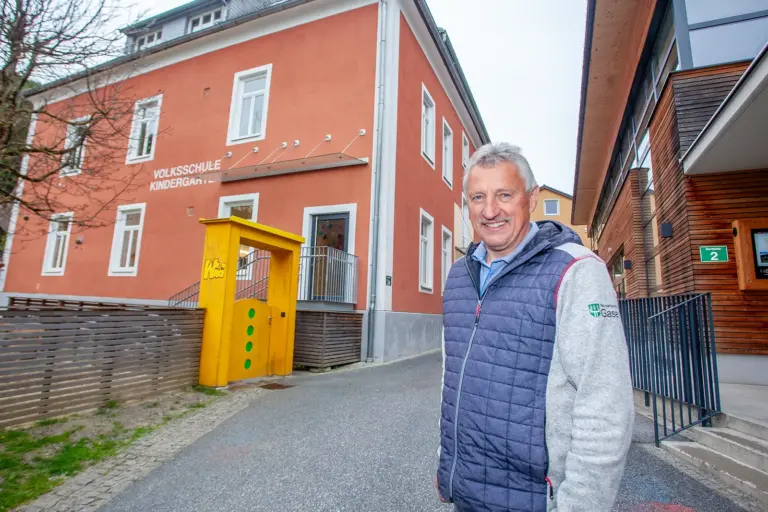 „Bis dahin Vollgas!“ – Bürgermeister von Gasen hört mit Jahreswechsel auf Ein Mann steht lächelnd auf der Straße, im Hintergrund links ein Haus mit der Aufschrift "Volksschule Kindergarten", rechts ein Haus mit Holzfassade