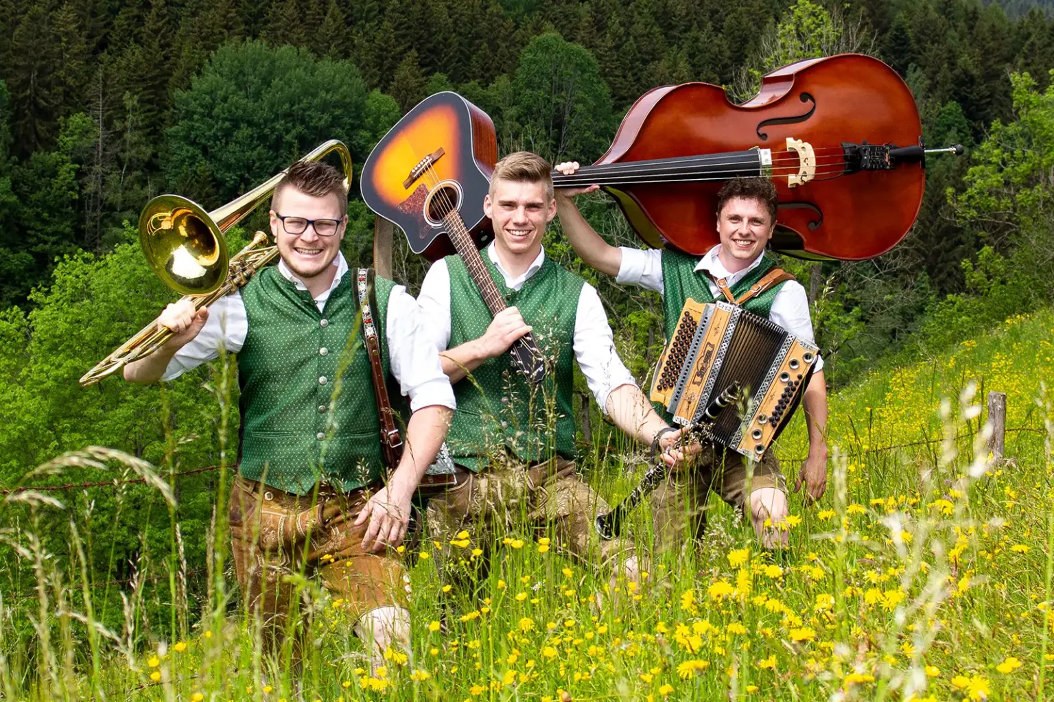 Die Goaßsteigbuam laden zum Fest Drei Musiker in Tracht stehen mit ihren Instrumenten auf einer blühenden Wiese vor einem Wald. Sie tragen Posaune, Gitarre, Kontrabass und Steirische Harmonika und lächeln in die Kamera.
