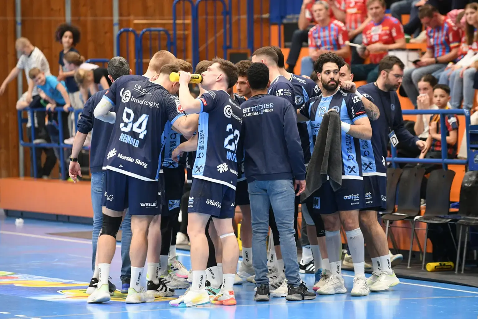 Spieler der BT Füchse versammeln sich während einer Spielunterbrechung in einem engen Kreis an der Seitenlinie der Handballhalle; einige trinken aus Trinkflaschen, während Trainer und Betreuer Anweisungen geben. Im Hintergrund sitzen Zuschauer auf der Tribüne.
