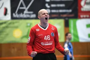 Ein Handballspieler im roten Trikot mit der Nummer 46 jubelt lautstark auf dem Spielfeld und ballt beide Fäuste. Im Hintergrund sind unscharf Werbebanner und ein weiterer Spieler in einer Sporthalle zu sehen.
