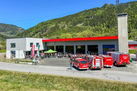 Ein Gebäude mit Turm und roter Fassade mit rotem Vordach, davor vier Feuerwehrfahrzeuge und zahlreiche Menschen, teils stehend, teils an Biertischen sitzend