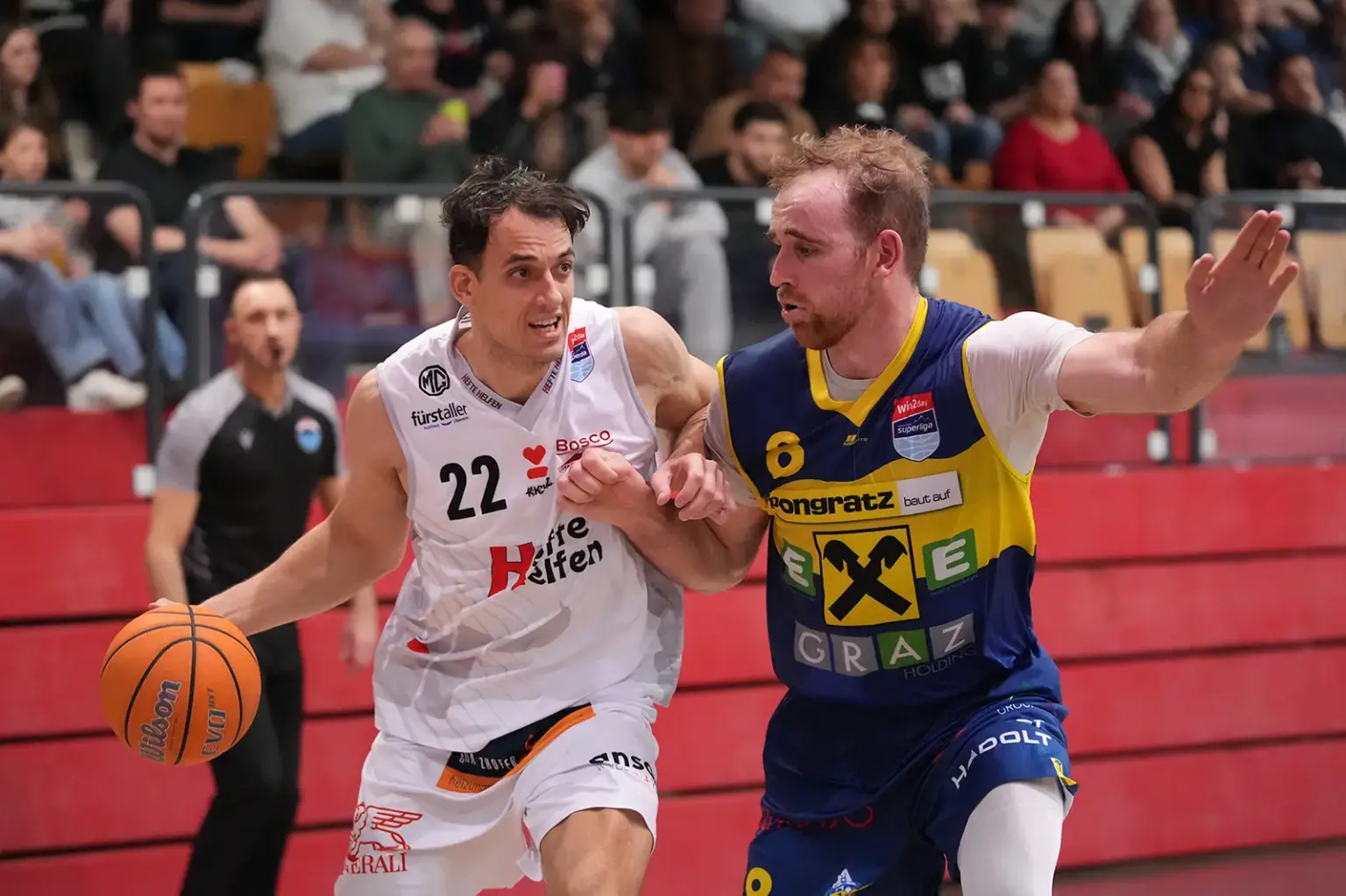 Zwei Basketballspieler kämpfen in einer Spielsituation um die bessere Position: Ein Spieler im weißen Kapfenberg-Trikot dribbelt mit dem Ball Richtung Korb, während ihn ein Gegenspieler im blau-gelben Dress eng verteidigt; im Hintergrund verfolgen Zuschauer das Spiel von den Tribünen.
