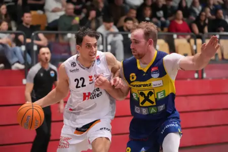 Zwei Basketballspieler kämpfen in einer Spielsituation um die bessere Position: Ein Spieler im weißen Kapfenberg-Trikot dribbelt mit dem Ball Richtung Korb, während ihn ein Gegenspieler im blau-gelben Dress eng verteidigt; im Hintergrund verfolgen Zuschauer das Spiel von den Tribünen.