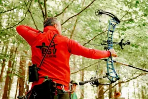 Ein Bogenschütze des BSV Kapfenberg spannt im Wald einen Compoundbogen und visiert konzentriert sein Ziel an. Der Sportler trägt eine rote Vereinsjacke und komplette Schießausrüstung.