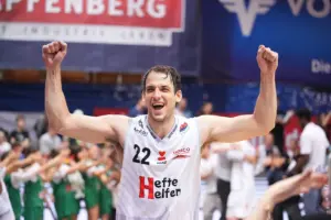 Jubel in der Basketballhalle: Ein Spieler im weißen Trikot reißt nach dem Spiel beide Arme in die Höhe, während Fans und Teamkollegen im Hintergrund mitfeiern.