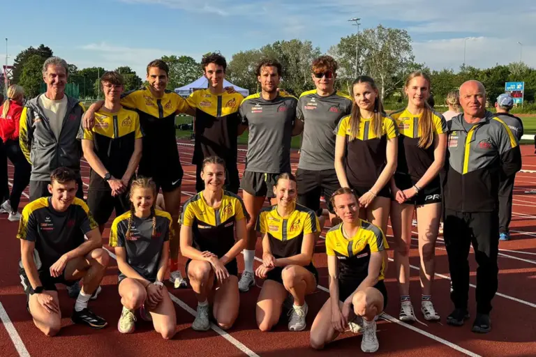 Steirischer Rekord beim Heimwettkampf Eine Gruppe von Athletinnen, Athleten und Trainern posiert in Vereinskleidung auf einer Laufbahn für ein gemeinsames Mannschaftsfoto. Im Hintergrund sind die Leichtathletikanlage, Bäume und ein sonniger Abendhimmel zu sehen.